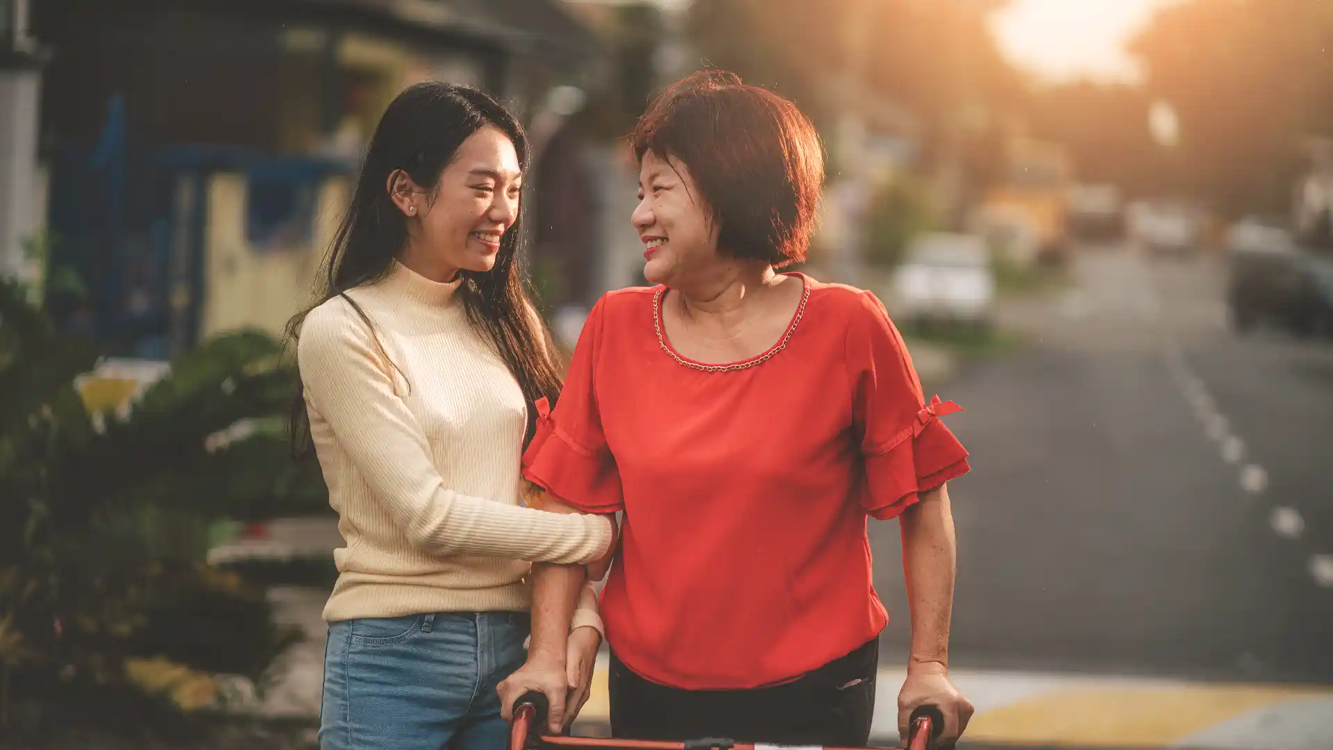 A smiling young woman supports the arm of a senior woman who is using a red walker on a residential street.